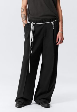Person wearing loose black trousers with a long white string belt, black shoes, a black top, and a tattoo visible on the left arm.