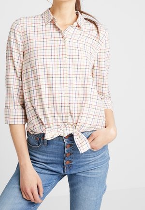 Kobieta ubrana w białą koszulę w kratę zapinaną na guziki, związana w talii, z podwiniętymi rękawami oraz w wysokiej talii niebieskie dżinsy z widocznymi guzikami.