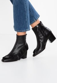 Bottes courtes en cuir noir avec un talon épais, ornées de volants et d'une fermeture éclair latérale, associées à un jean bleu raccourci.