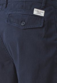 Mornarsko plave pamučne chinos hlače s stražnjim džepom s dugmetom, teksturirana tkanina i vidljiva oznaka s natpisom "Guess Jeans."