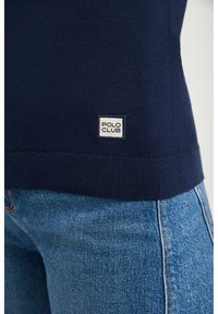 Marinblå tröja med "POLO CLUB"-etikett i nederkant, ihop med ljusblå denimjeans, med fokus på midje- och höftområdet.