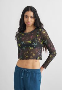 Γυναίκα με μακριά σκούρα μαλλιά που φοράει μαύρο διαφανές crop top με μακριά μανίκια και πολύχρωμα σχέδια αστερίας, καθώς και μπλε παντελόνι με κορδόνι στη μέση.