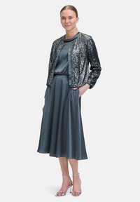 Silberne Paillettenjacke über einem dunkelblauen Satin-Kleid mit schwingendem Saum, das kurze Ärmel und eine lockere Passform hat. Kombiniert mit silbernen, verzierten Sandalen.