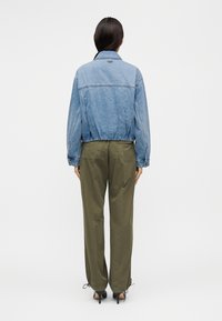 Τζιν μπουφάν με ελαστικό κάτω μέρος, ανοιχτό μπλε χρώμα· συνδυασμένο με παντελόνι σε ελιά πράσινη απόχρωση, με δύο πλαϊνές τσέπες και ρυθμιζόμενες ραφές στο τελείωμα.