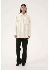 Crème knoopshirt met twee borstzakken, lange mouwen en een gestructureerde stof, gecombineerd met zwarte broeken en puntige hakken.