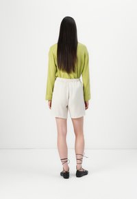Groene geribde lange mouwen shirt, beige elastische taille shorts, zwarte veterschoenen. Model staat met de rug naar de camera tegen een witte achtergrond.