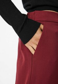 Bordeauxkleurige broek met een gladde textuur, voorzien van een zijzak. Een zwarte trui met geribbelde boorden is gedeeltelijk zichtbaar.