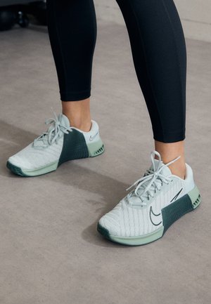 Person trägt weiße und grüne Nike-Sneaker mit schwarzen Leggings und steht auf einem glatten beigen Boden.
