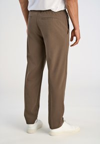 Braune, taillierte Hose mit lockerer Passform, elastischem Bund und Gesäßtaschen, kombiniert mit weißen Sneakers vor einem hellen Hintergrund.