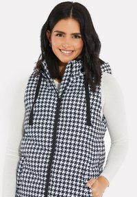 Kvadra-stickad puffer väst i svart och vit hundtandsmönster, med hög krage, dragkedja fram och justerbara snoddar vid huven.
