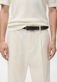 Kremowy sweter z dzianiny zestawiony z jasnobeżowymi spodniami. Ciemnobrązowy skórzany pasek z srebrną klamrą podkreśla strój. Konstrukcja z płaskim przodem.