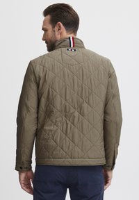 Quilted olive green jacket with a stand collar, featuring red, navy, and white striped accents at the neck and black snap buttons on the sleeves.