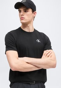 Νέος άνδρας που φοράει μαύρο μπλουζάκι Calvin Klein και μαύρο καπέλο, με σταυρωμένα χέρια, κοιτάζοντας πλαγίως σε απλό φωτεινό φόντο.