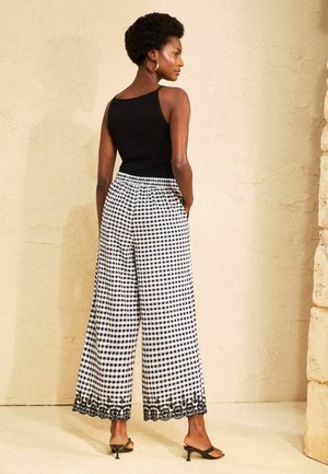 Žena stojaca zboku má na sebe čierny tielko bez rukávov, široké čierno-biele kárované nohavice s čipkovaným lemom a čierne sandále na podpätku.