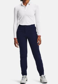 Polo blanc à manches longues avec patte de boutonnage, associé à un pantalon bleu marine à coupe slim avec des accents sur les côtés. Les chaussures sont gris clair avec des lacets.