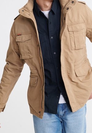 Mann trägt eine hellbraune Jacke mit mehreren Vordertaschen über einem schwarzen Hemd mit Knöpfen und hellblauen Jeans, vor einem einfarbigen Hintergrund.