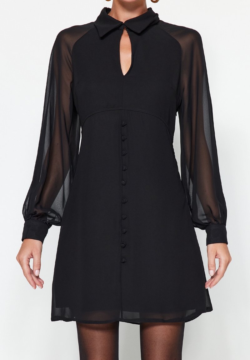 Schwarzes Kleid mit durchsichtigen langen Ärmeln, einem Ausschnitt mit Schlüsselöffnung und Knopfdetails auf der Vorderseite. Leichte, fließende Stoffqualität mit strukturierter Silhouette.