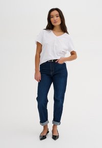 Biela tričko s výstrihom do V, modré riflové nohavice s manžetami a čierne topánky so špicatou špičkou. Hladká tkanina a uvoľnený strih.