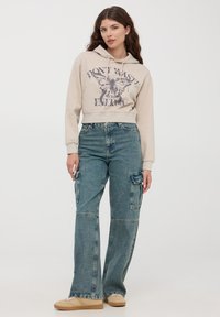 Beżowa, skrócona bluza z kapturem z graficznym nadrukiem, w połączeniu z luźnymi, wyblakłymi niebieskimi spodniami cargo z kieszeniami z przodu i z boku. Beżowe trampki dopełniają całość.