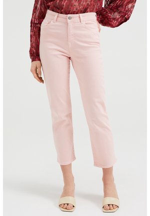 Vrouw die lichtroze cropped jeans, een rood gebloemde blouse en beige sandalen met open tenen draagt, staand tegen een effen achtergrond.