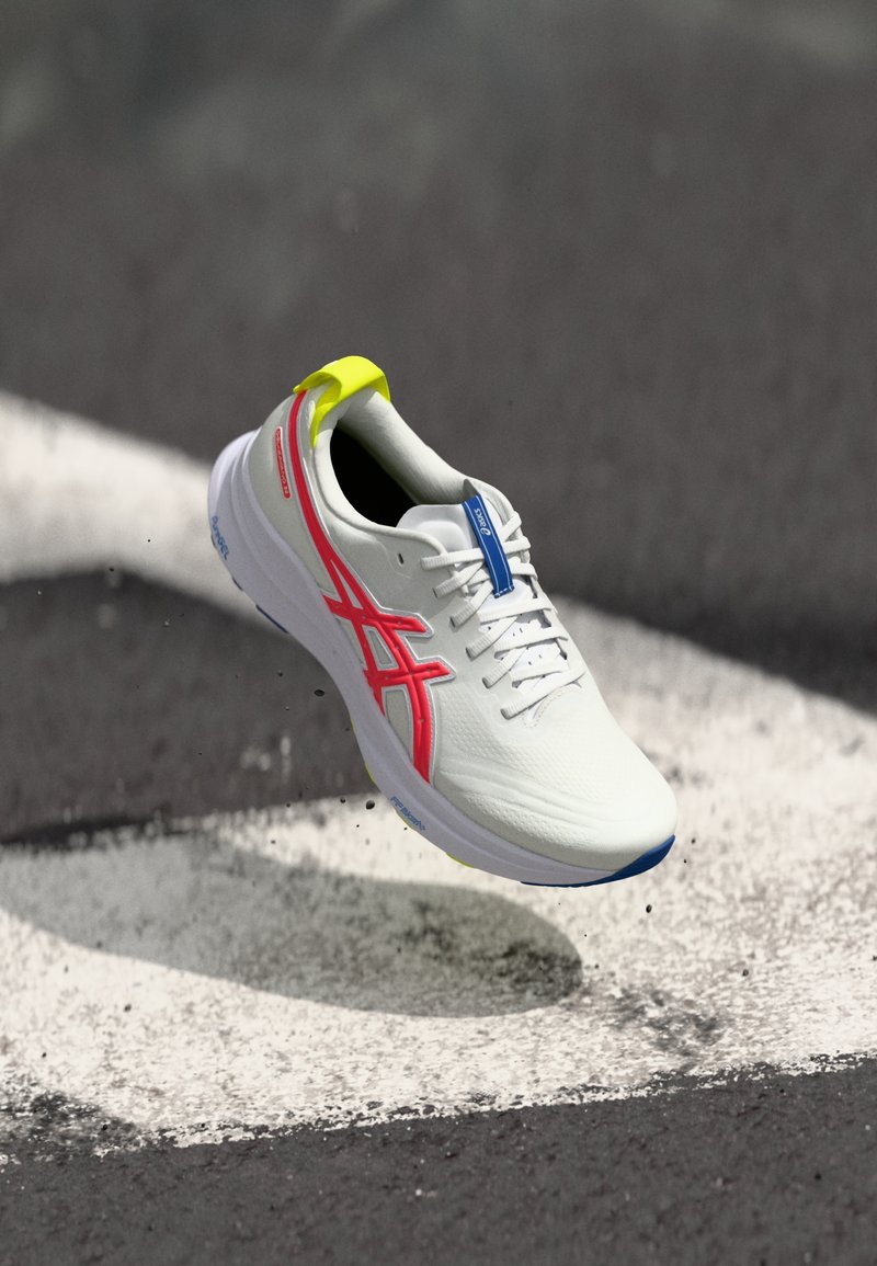 White athletic shoe with pink and blue accents, textured upper, yellow heel tab, and visible lacing system, suspended above a mark on pavement.