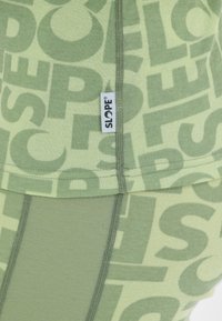 Groene gelaagde top met een herhaald tekstpatroon in lichter groen. Het materiaal is zacht met zichtbare stiksels. Merktag met het label "SLOPE."