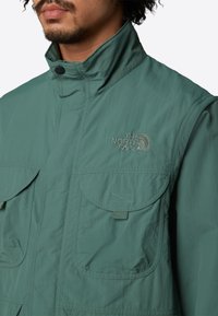 Grüne Nylonjacke mit hohem Kragen, zwei großen Fronttaschen und gesticktem Logo auf der Brust. Glatte Textur und Outdoor-Design.