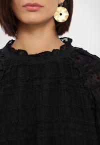 Top nero con texture e colletto ondulato, caratterizzato da tessuto a strati e accenti a pois. Orecchini circolari dorati con una perlina nera indossati.