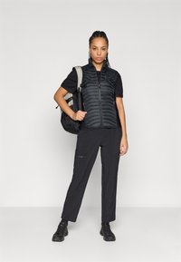 Černá zateplená vesta se zipem vpředu, v kombinaci s černým tričkem a cargo kalhotami. Oblečení doplňuje šedý batoh a černé boty.