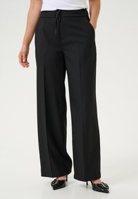 Pantaloni negri cu pantaloni largi, cu un cordon la talie, buzunare laterale și o textură netedă, asortați cu tocuri negre cu vârful ascuțit.
