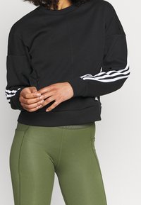 Felpa nera con maniche lunghe, caratterizzata da strisce bianche, tessuto a trama, scollo rotondo e vita aderente, abbinata a leggings verde oliva.