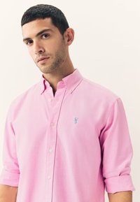Light pink button-up shirt with a collared neck and rolled sleeves. Features a small blue emblem on the chest. Smooth, cotton fabric.