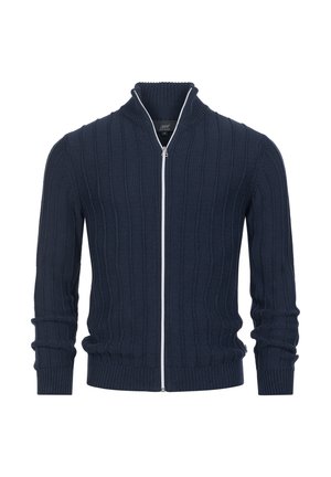 Marineblå genser med glidelås og høy krage, med strukturert ribbestrikk, lange ermer og en kontrasterende hvit glidelås. Stoffet ser ut til å være strikket.