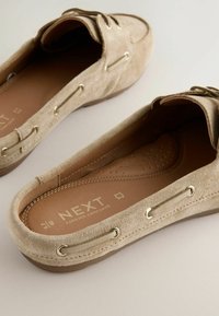 Beige suede slip-on shoes with metal eyelets, textured inner lining, and a flexible rubber sole, featuring embossed branding on the insole.