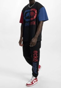 Zwarte t-shirt met rode en blauwe mouwen, voorzien van een logo en tekst in contrasterende kleuren; zwarte joggers met rode en blauwe tekstaccenten; rode sneakers.