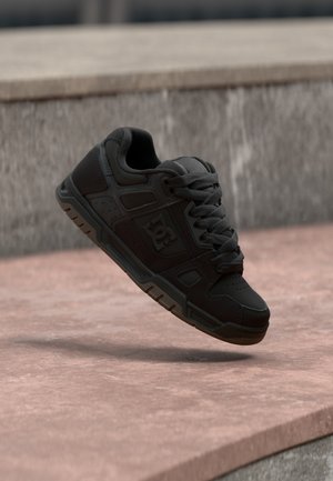 Schwarzer Skate-Schuh mit Schnürsenkeln in der Luft über einer pinken Betonoberfläche und grauen Steinstufen im Hintergrund.
