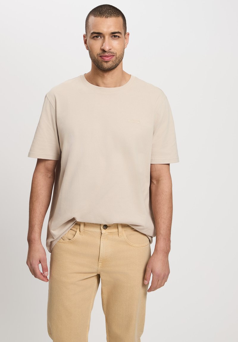 Beżowa bawełniana koszulka z krótkim rękawem i okrągłym dekoltem, w zestawieniu z jasnobeżowymi dżinsami, z delikatnym logo na piersi.