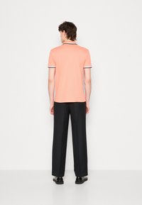 Herren Polo-Shirt mit kurzen Ärmeln in Koralle, mit kontrastierendem schwarzem Kragen und Ärmelbesatz, kombiniert mit schwarzen maßgeschneiderten Hosen und Schuhen.