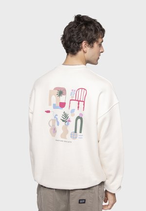 Sweatshirt color crema con una grafica multicolore che presenta forme astratte, piante e una sedia sul retro. Design a vestibilità comoda.