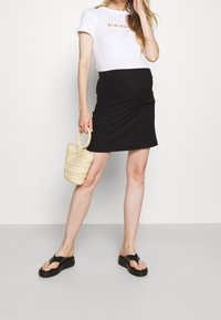 Femme portant un t-shirt blanc "Je ne sais", une jupe de maternité noire, des sandales à plateforme noires, tenant un petit sac à main en osier, se tenant devant un fond blanc.