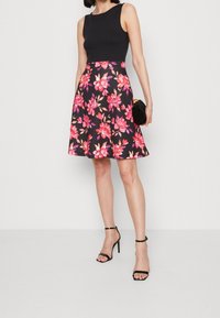 Top sans manches noir associé à une jupe florale longueur genou comportant des fleurs roses et oranges sur un fond noir. Accessoirisé avec un petit sac à main noir et des talons aiguilles noirs.