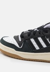 Närbild av svart, vit och mörkgrön sneaker med perforerat tåkappa, svarta snörningar, tre svarta ränder och en beige sula.