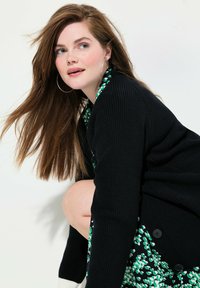 Cardigan noir à côtes avec de grands boutons, porté sur une robe à motif floral vert et noir. De longs cheveux bruns tombent librement.
