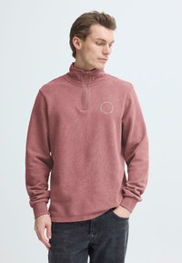 Trui in een gedempte rozekleur, voorzien van een halve rits bij de hals, geribbelde boorden en een cirkelvormig logo op de linkerborst. Zachte textuur.