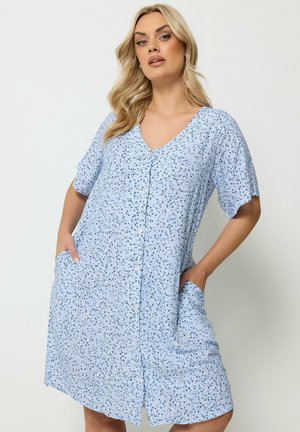 Lyshåret kvinde iført en lyseblå kjole med korte ærmer og små blomsterprint, hænderne i lommerne, stående mod en hvid baggrund.