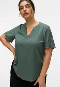 Blusa de manga corta en verde con escote en V, confeccionada en tela suave. Presenta un corte holgado y un dobladillo ligeramente curvado. Sin patrones visibles ni detalles de herrajes.