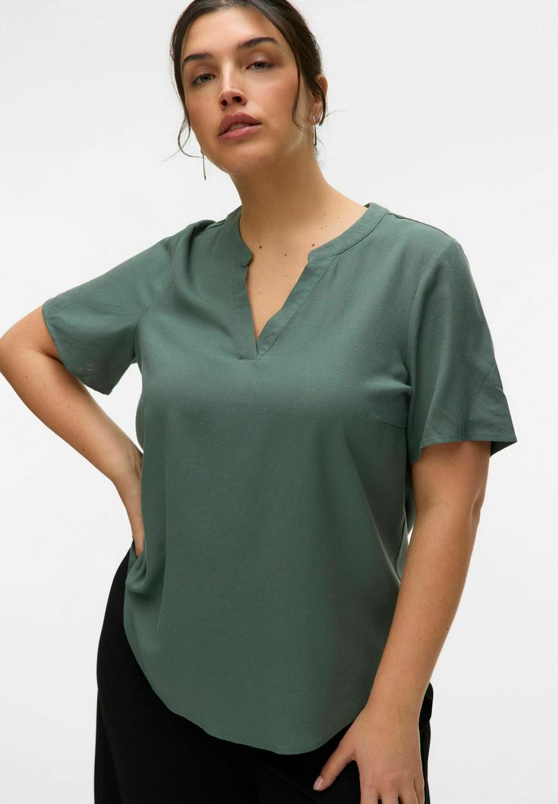 Blusa de manga corta en verde con escote en V, confeccionada en tela suave. Presenta un corte holgado y un dobladillo ligeramente curvado. Sin patrones visibles ni detalles de herrajes.