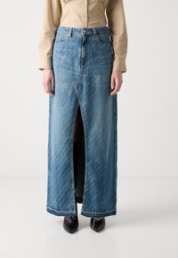 Kvinna klädd i en beige blus och en lång blå denimkjol med en framklippning och fransar i fållen, tillsammans med svarta spetsiga stövlar.