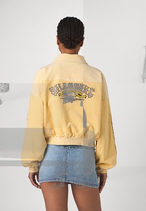 Femme aux cheveux tressés portant une veste Billabong jaune et une mini-jupe en jean bleue, vue de dos sur un fond uni.