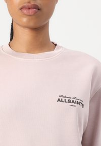 Prim-plan cu o persoană care poartă un pulover roz deschis cu textul „Infinite Summer ALLSAINTS LONDON” imprimat pe piept.